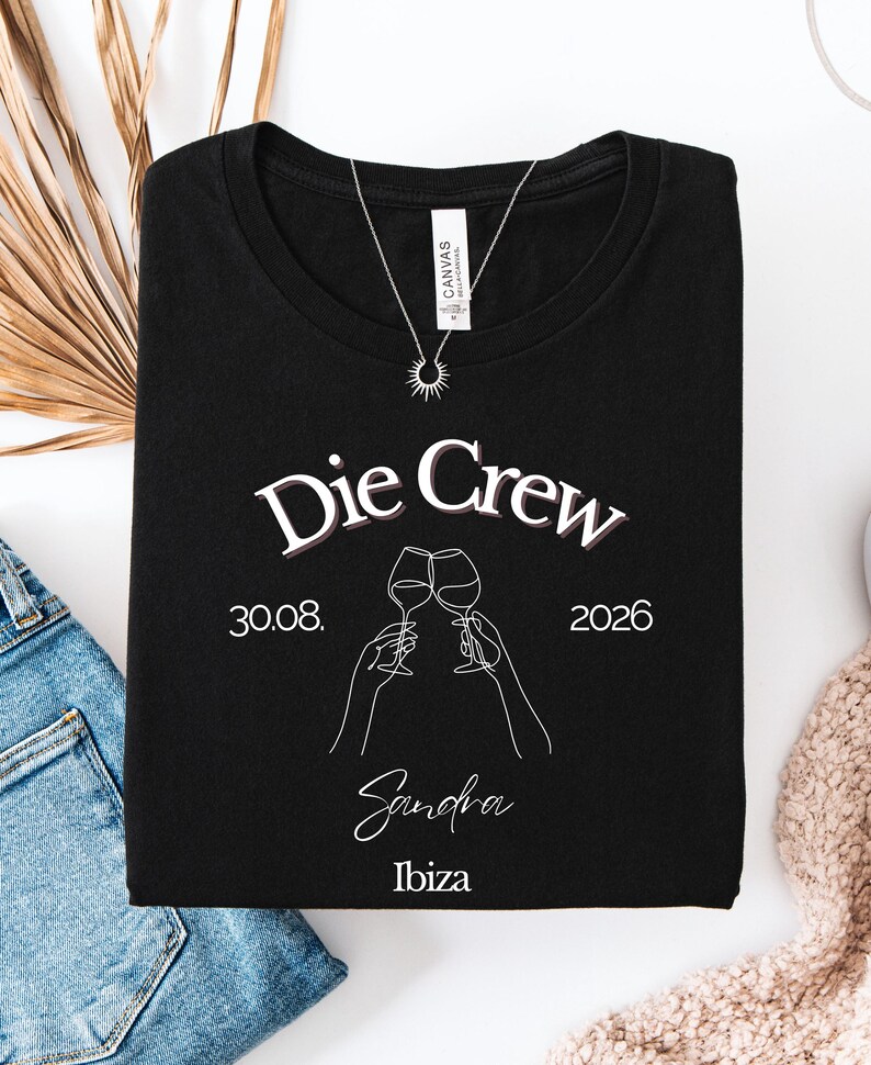 May include: A black t-shirt featuring the white text Die Crew, the date 30.08. 2026, the name Sandra, and the word Ibiza. The graphic includes a white line art illustration of two hands holding wine glasses in a toast, centered on the chest.