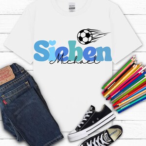 May include: White t-shirt with the name "Sieben Michael" in blue and black, a soccer ball graphic, and a pair of black Converse sneakers. Dark blue jeans and a bundle of colored pencils are also in the image.