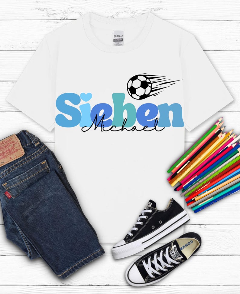 May include: White t-shirt with the name "Sieben Michael" in blue and black, a soccer ball graphic, and a pair of black Converse sneakers. Dark blue jeans and a bundle of colored pencils are also in the image.