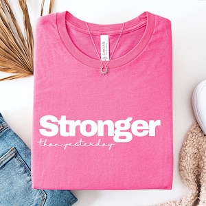 May include: A vibrant pink t-shirt featuring the text "Stronger than yesterday" in white. A silver necklace with a sunburst pendant is placed on the shirt. The shirt is folded, with blue jeans and a beige scarf in view.