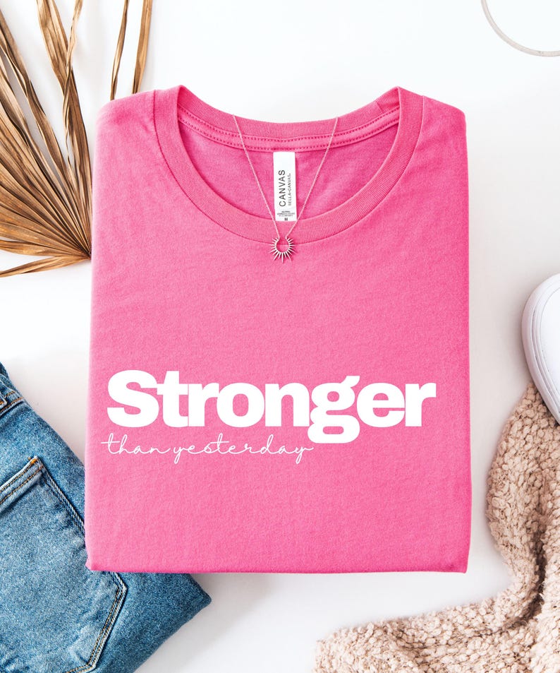 May include: A vibrant pink t-shirt featuring the text "Stronger than yesterday" in white. A silver necklace with a sunburst pendant is placed on the shirt. The shirt is folded, with blue jeans and a beige scarf in view.