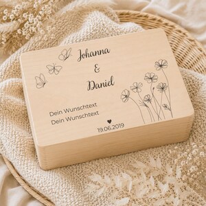 May include: A rectangular light-colored wooden box featuring black laser-engraved designs including three butterflies, a floral arrangement, and the text Johanna & Daniel, Dein Wunschtext, Dein Wunschtext, and the date 19.06.2019 with a small heart.