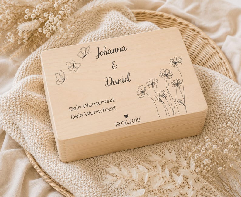 May include: A rectangular light-colored wooden box featuring black laser-engraved designs including three butterflies, a floral arrangement, and the text Johanna & Daniel, Dein Wunschtext, Dein Wunschtext, and the date 19.06.2019 with a small heart.