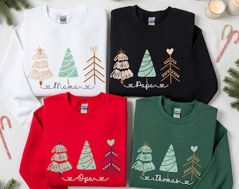 Puede incluir: Cuatro sudaderas de cuello redondo en blanco, negro, rojo y verde. Cada una presenta un diseño de árbol de Navidad con las palabras "Mama", "Papa", "Opa" y "Thomas". Las sudaderas tienen un tema festivo navideño.