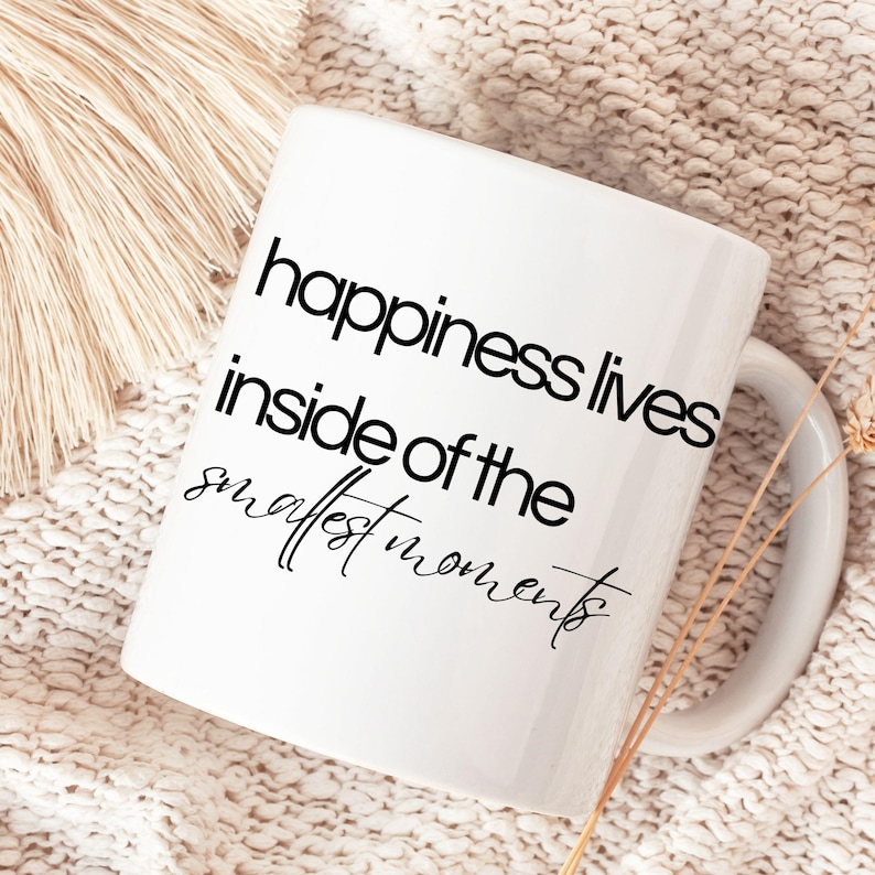 Może przedstawiać: Biały ceramiczny kubek z zakrzywionym uchwytem. Kubek ma napis "happiness lives inside of the smallest moments" czarnymi literami. Kubek jest umieszczony na tle o neutralnej fakturze.