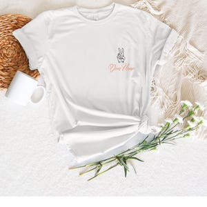 May include: White t-shirt with a peace sign graphic and the text "Dun Name" in a cursive font. The shirt is knotted at the waist. A white mug and a woven basket are in the background.