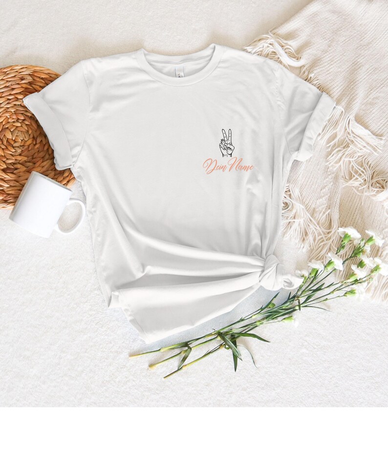 May include: White t-shirt with a peace sign graphic and the text "Dun Name" in a cursive font. The shirt is knotted at the waist. A white mug and a woven basket are in the background.