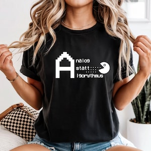 May include: Black t-shirt with white pixelated letter "A" and text "analog statt: 1980rythmus" and a Pac-Man-like character. The shirt is a classic crew neck style.