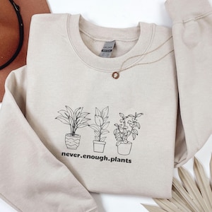 Könnte beinhalten: Beiges Sweatshirt mit Rundhalsausschnitt und langen Ärmeln. Auf der Vorderseite befindet sich eine schwarze Strichzeichnung von drei Topfpflanzen und der Text "never.enough.plants". Eine goldene Halskette mit Anhänger ist sichtbar.