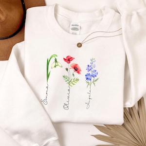Puede incluir: Sudadera blanca con diseños florales en acuarela y los nombres Emma, Olivia y Angela. Las flores incluyen un galanto, amapolas rojas y delfinios azules. Un collar dorado está colocado sobre la sudadera.