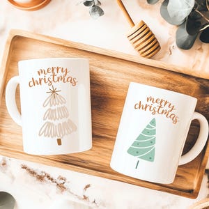 Op de afbeelding: Twee witte keramische mokken met de woorden "merry christmas" in koperkleurig schrift. Eén mok heeft een lichtbruin boomontwerp en de andere een groen boomontwerp. De mokken staan op een houten dienblad.