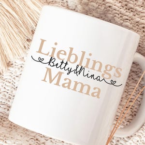 Puede incluir: Taza de cerámica blanca con las palabras "Lieblings Mama Betty & Nina" en una mezcla de escritura marrón y negra. La taza tiene un asa curva y está sobre un fondo neutro y texturizado.