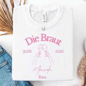 May include: A white t-shirt featuring pink text that reads Die Braut, 30.08. 2026, Manuela, and Ibiza. The design includes a line art illustration of two hands holding wine glasses in a toast. A silver necklace with a sun pendant rests on the collar.