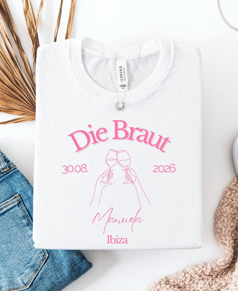 May include: A white t-shirt featuring pink text that reads Die Braut, 30.08. 2026, Manuela, and Ibiza. The design includes a line art illustration of two hands holding wine glasses in a toast. A silver necklace with a sun pendant rests on the collar.