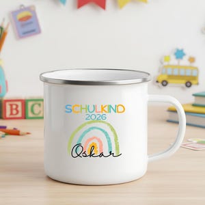 May include: A white enamel mug with a silver rim featuring a colorful rainbow graphic, the text "SCHULKIND 2026" in blue and orange, and the name "Oskar" written in black cursive script across the front of the mug.