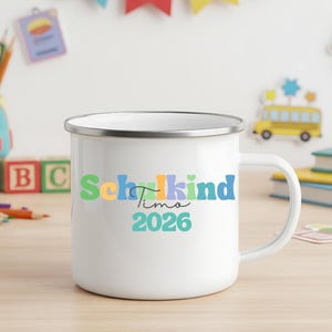 May include: A white enamel mug with a silver rim featuring the colourful text Schulkind, the name Timo in black script, and the year 2026 in teal. The mug has a side handle and a smooth finish, displaying a vibrant design on the front surface.