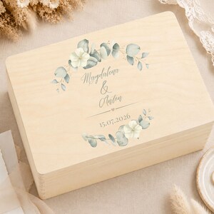 May include: A rectangular light-colored wooden box featuring a printed design of green eucalyptus leaves and white flowers. The text "Magdalena & Anton" is centered between the floral elements, followed by a small heart and the date "15.07.2026".