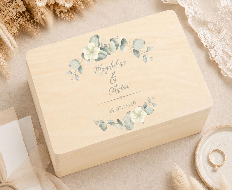 May include: A rectangular light-colored wooden box featuring a printed design of green eucalyptus leaves and white flowers. The text "Magdalena & Anton" is centered between the floral elements, followed by a small heart and the date "15.07.2026".