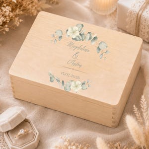May include: A rectangular light-colored wooden box featuring a printed design of green eucalyptus leaves and white flowers. The text "Magdalena & Anton" is printed in the center, followed by the date "15.07.2026" below a small heart icon.