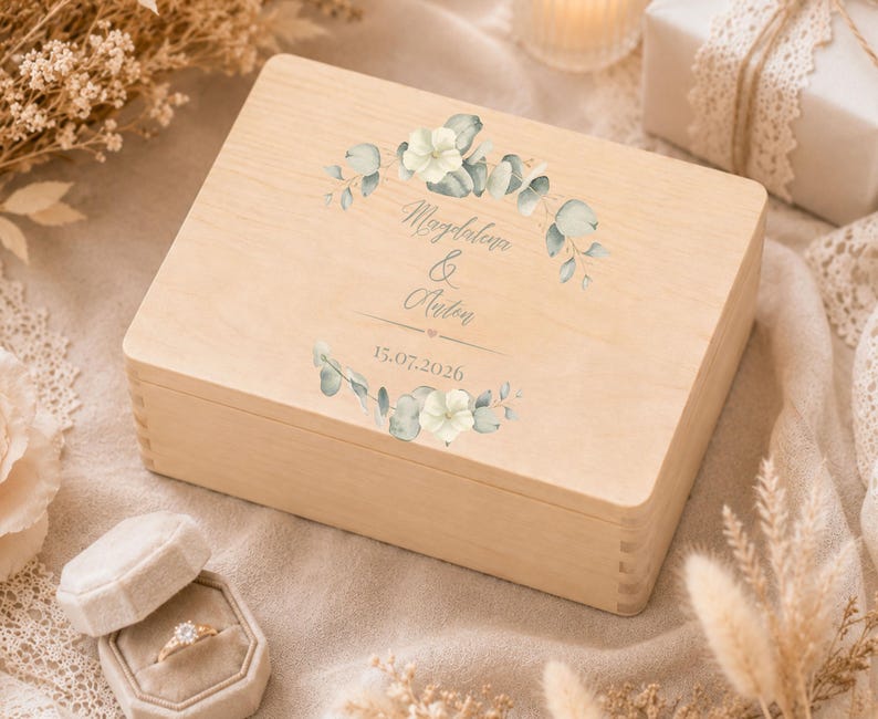 May include: A rectangular light-colored wooden box featuring a printed design of green eucalyptus leaves and white flowers. The text "Magdalena & Anton" is printed in the center, followed by the date "15.07.2026" below a small heart icon.