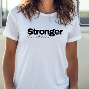 May include: A white t-shirt featuring the text "Stronger than yesterday" in black. The word "Stronger" is in a bold font, with "than yesterday" in a script style. The shirt has short sleeves and a crew neck.