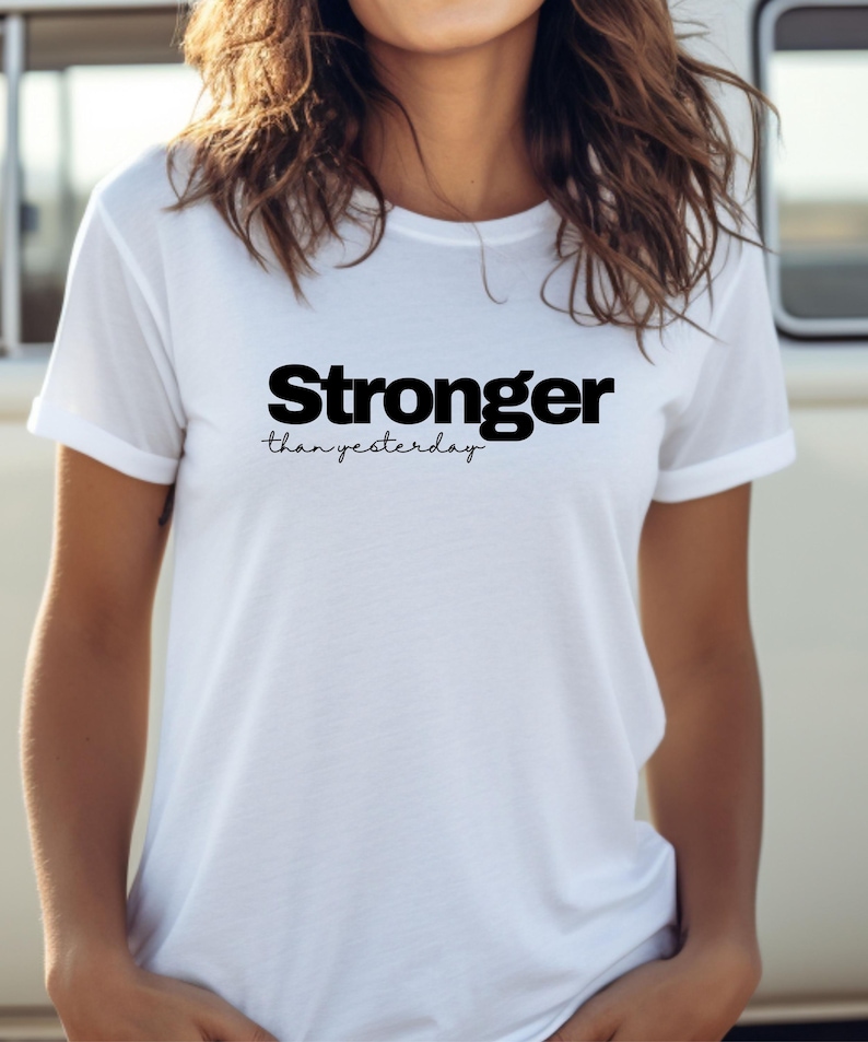 May include: A white t-shirt featuring the text "Stronger than yesterday" in black. The word "Stronger" is in a bold font, with "than yesterday" in a script style. The shirt has short sleeves and a crew neck.