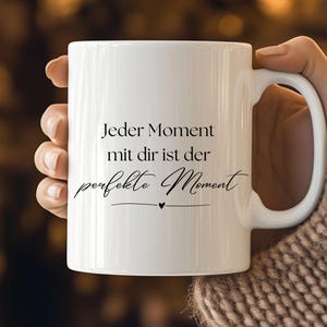 Puede incluir: Taza de cerámica blanca con asa curva. La taza presenta el texto "Jeder Moment mit dir ist der perfekte Moment" en escritura negra, con un pequeño símbolo de corazón. La taza está sujeta por una persona.