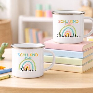 May include: Two white enamel mugs with silver rims, each featuring a colorful rainbow graphic, the text SCHULKIND 2026, and a personalized name in black script. One mug is labeled Charlotte and the other is labeled Oskar, both displayed on books.