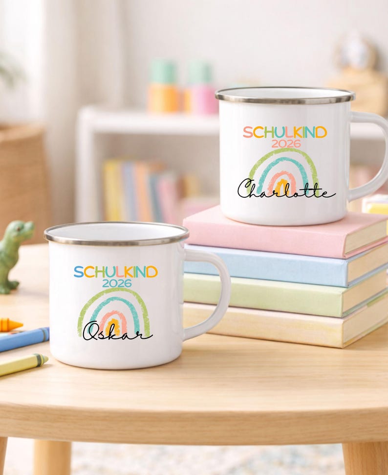 May include: Two white enamel mugs with silver rims, each featuring a colorful rainbow graphic, the text SCHULKIND 2026, and a personalized name in black script. One mug is labeled Charlotte and the other is labeled Oskar, both displayed on books.