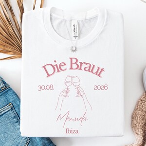 May include: A white t-shirt featuring pink text that reads Die Braut, 30.08. 2026, Manuela, and Ibiza. The design includes a line art illustration of two hands holding wine glasses in a toast. The shirt has a crew neckline and a short-sleeved cut.