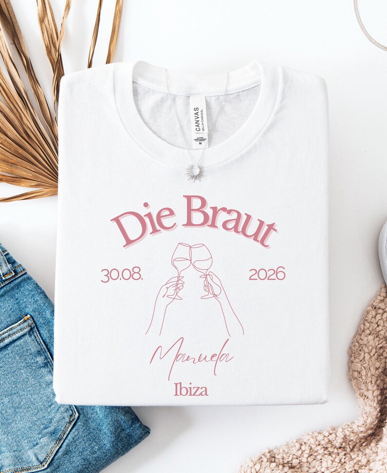 May include: A white t-shirt featuring pink text that reads Die Braut, 30.08. 2026, Manuela, and Ibiza. The design includes a line art illustration of two hands holding wine glasses in a toast. The shirt has a crew neckline and a short-sleeved cut.