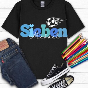 May include: Black t-shirt with the name "Sieben" in blue and a soccer ball graphic. The name "Michael" is written below in cursive. The shirt is paired with blue jeans and black Converse sneakers. Colored pencils are also in the image.