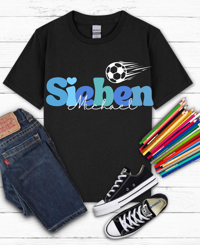 May include: Black t-shirt with the name "Sieben" in blue and a soccer ball graphic. The name "Michael" is written below in cursive. The shirt is paired with blue jeans and black Converse sneakers. Colored pencils are also in the image.