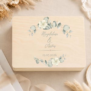 May include: A rectangular light-colored wooden box featuring a printed design of green eucalyptus leaves and white flowers arranged in a wreath. The text "Magdalena & Anton" is centered, followed by a small heart and the date "15.07.2026".