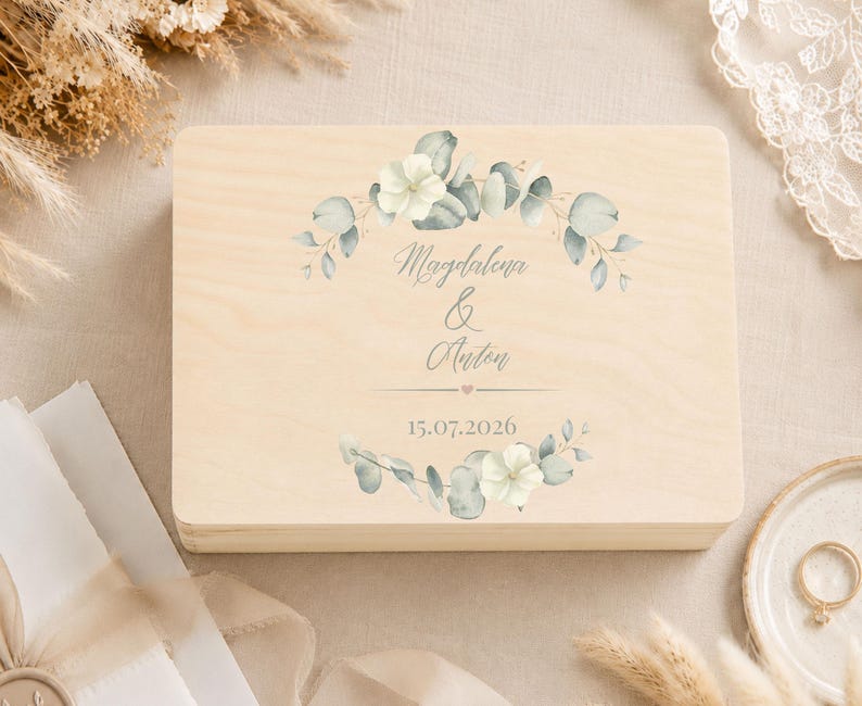 May include: A rectangular light-colored wooden box featuring a printed design of green eucalyptus leaves and white flowers arranged in a wreath. The text "Magdalena & Anton" is centered, followed by a small heart and the date "15.07.2026".