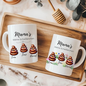 Puede incluir: Dos tazas de cerámica blanca con el texto "Mama's kleine Scheißerchen" y emojis de caca de dibujos animados. Una taza tiene tres emojis con nombres debajo, la otra tiene los mismos con flores. Las tazas están en una bandeja de madera.