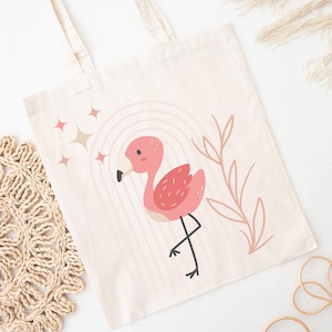 May include: A cream-colored tote bag featuring a pink flamingo illustration with a rainbow and star accents. The flamingo has a black beak and is standing on one leg. The bag has two handles.