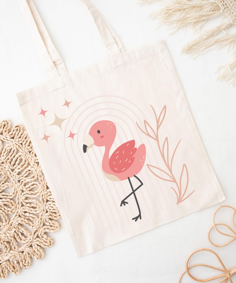 May include: A cream-colored tote bag featuring a pink flamingo illustration with a rainbow and star accents. The flamingo has a black beak and is standing on one leg. The bag has two handles.