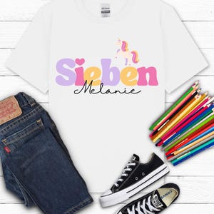 K&ouml;nnte beinhalten: Wei&szlig;es T-Shirt mit dem Namen "Sieben Melanie" in Lila, Gelb und Rosa und einer Einhorn-Grafik. Das Shirt wird mit blauen Jeans und schwarzen Converse-Sneakern kombiniert. Ein B&uuml;ndel Buntstifte ist ebenfalls vorhanden.