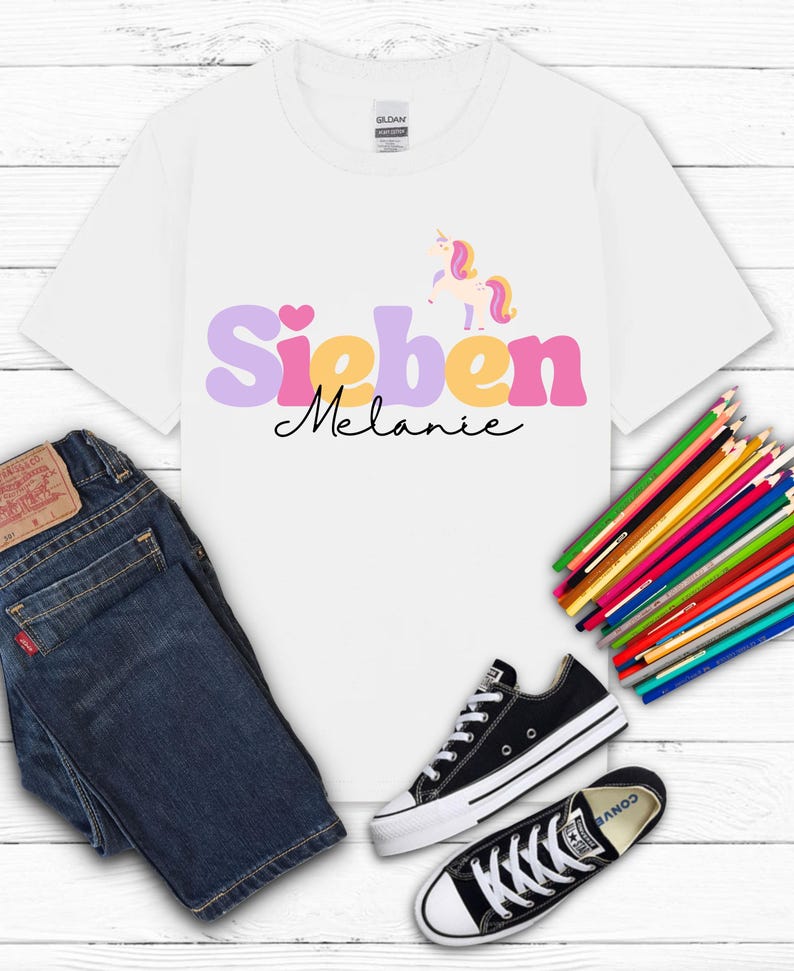K&ouml;nnte beinhalten: Wei&szlig;es T-Shirt mit dem Namen "Sieben Melanie" in Lila, Gelb und Rosa und einer Einhorn-Grafik. Das Shirt wird mit blauen Jeans und schwarzen Converse-Sneakern kombiniert. Ein B&uuml;ndel Buntstifte ist ebenfalls vorhanden.