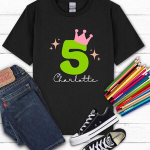 May include: Black t-shirt with a large green number 5, topped with a pink crown, and the name "Charlotte" in white script. The shirt is paired with blue jeans and black Converse sneakers. Colored pencils are arranged on a white wooden surface.