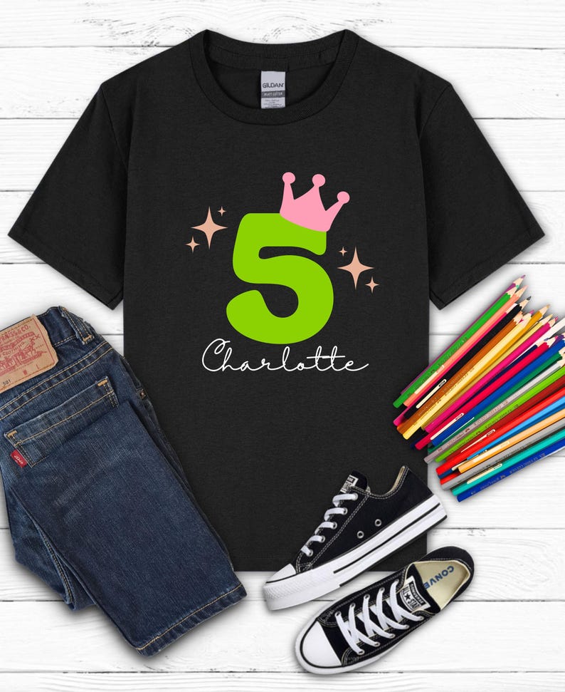 May include: Black t-shirt with a large green number 5, topped with a pink crown, and the name "Charlotte" in white script. The shirt is paired with blue jeans and black Converse sneakers. Colored pencils are arranged on a white wooden surface.