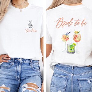 May include: White t-shirt with a peace sign and the text "Dear Name" on the front. The back of the shirt features the text "Bride to be" and illustrations of cocktails. The shirt is paired with blue jeans.