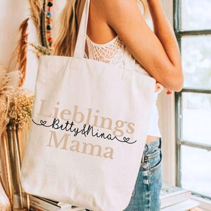 May include: Cream-colored tote bag with the words "Lieblings Betty & Nina Mama" printed in a mix of black and tan fonts. The bag has long straps and is being held by a person.