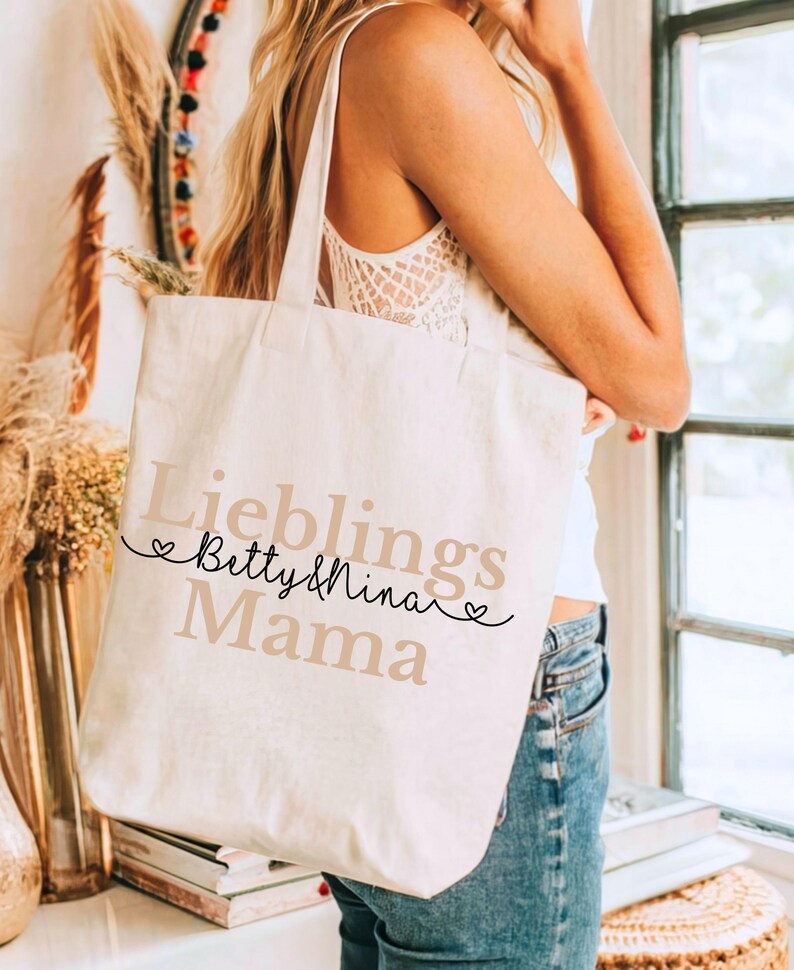 May include: Cream-colored tote bag with the words "Lieblings Betty & Nina Mama" printed in a mix of black and tan fonts. The bag has long straps and is being held by a person.