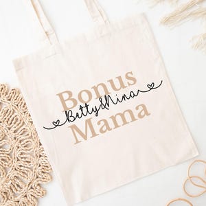 May include: Cream-colored tote bag with the words "Bonus Betty & Nina Mama" printed in a mix of serif and script fonts. The words are in shades of brown and black, with small heart accents. The bag has two handles.