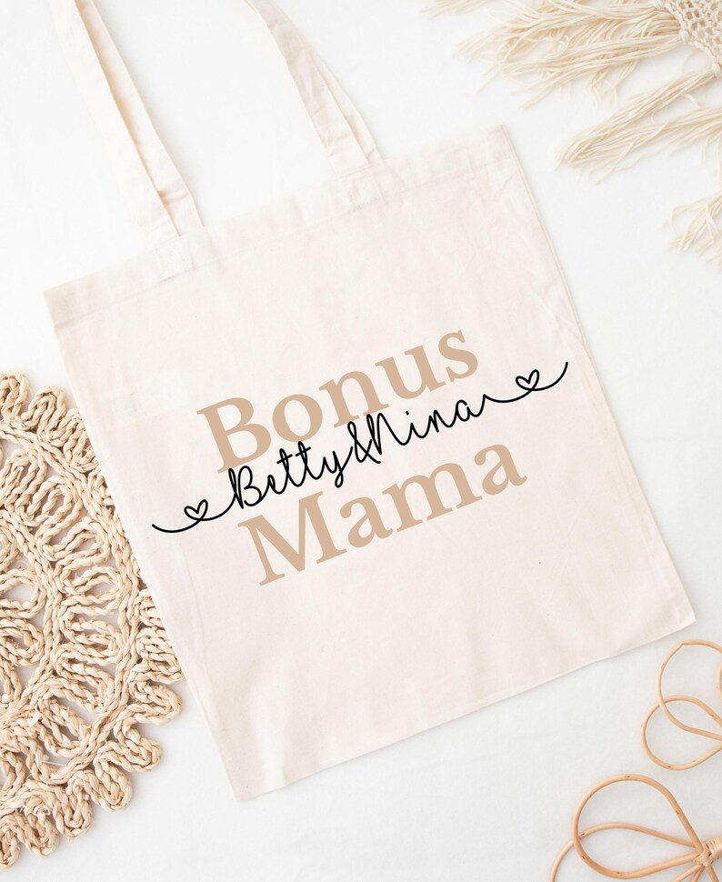 May include: Cream-colored tote bag with the words "Bonus Betty & Nina Mama" printed in a mix of serif and script fonts. The words are in shades of brown and black, with small heart accents. The bag has two handles.