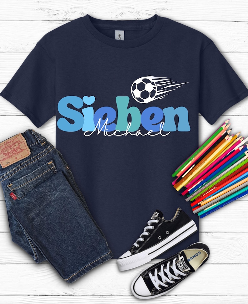 May include: Navy blue t-shirt with the name "Sieben Michael" in blue and a soccer ball graphic. The shirt is paired with blue jeans, black sneakers, and colored pencils on a white wooden surface.
