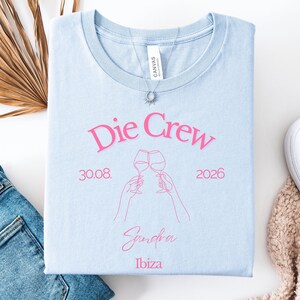 May include: A light blue t-shirt featuring pink text that reads Die Crew, 30.08. 2026, Sandra, and Ibiza. The design includes a pink line art illustration of two hands holding wine glasses in a toast. A silver sunburst pendant necklace rests on the collar.