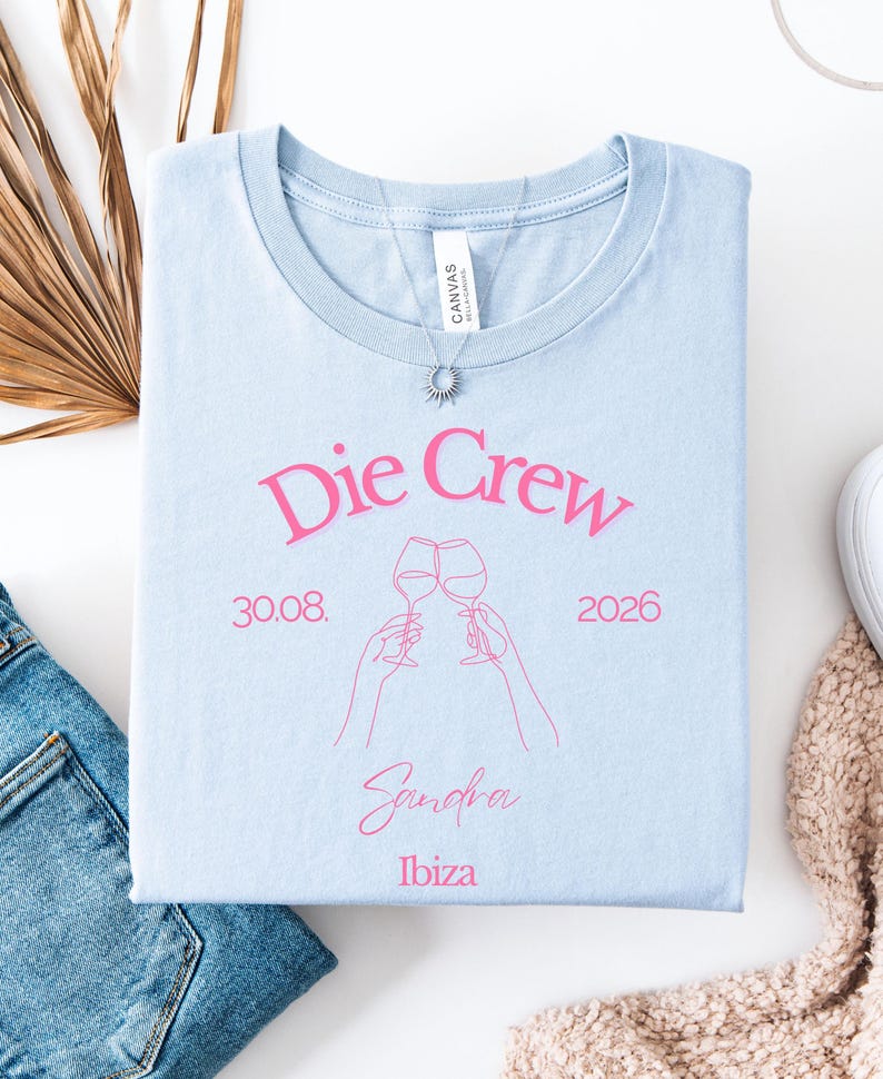 May include: A light blue t-shirt featuring pink text that reads Die Crew, 30.08. 2026, Sandra, and Ibiza. The design includes a pink line art illustration of two hands holding wine glasses in a toast. A silver sunburst pendant necklace rests on the collar.